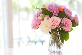 Bouquet of roses, snapdragons, chrysanthemums, and greenery in pink ruffled vase on tabler in front of sunlit window.