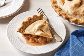 slice of apple pie on white plate with fork, whole pie and blue napkin nearby