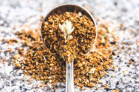 An antique spoon full of dukkah, a spice mix used in middle eastern cooking, spills out onto a stone table top